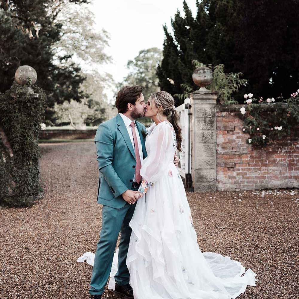 Groom in blue suit and pink tie kisses bride in long sleeve Victoria Sanders wedding dress with embroidery and train in the grounds of Iscoyd Park