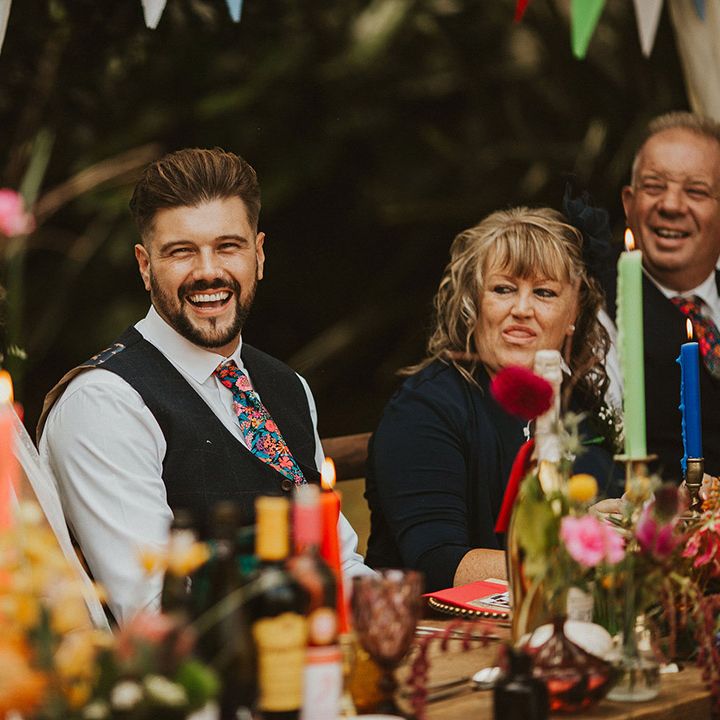 Groom in a navy blue waistcoat and floral Liberty print tie laughing during the clear marquee wedding reception speeches 