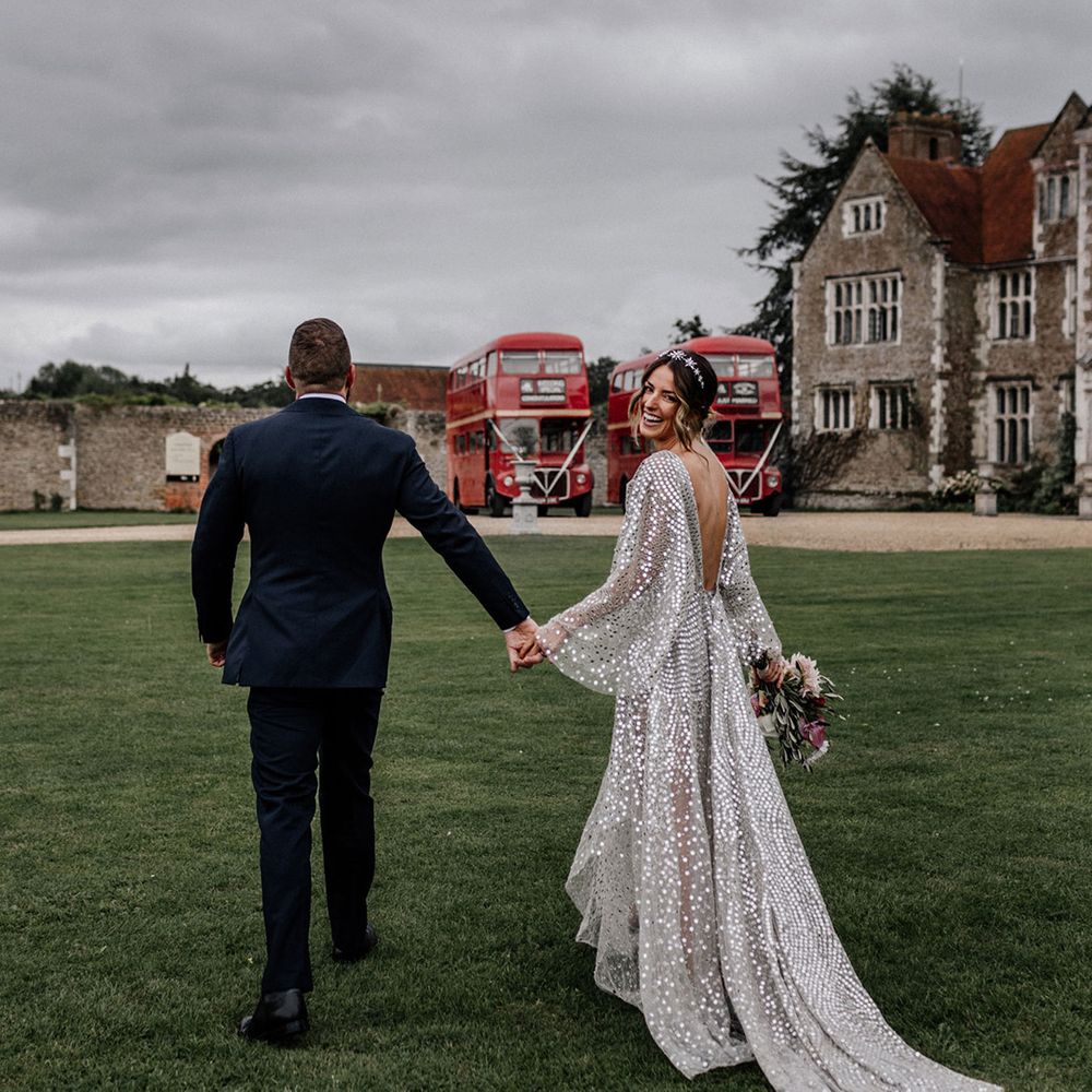 Groom in navy blue suit holding hands with his bride in a silver disc wedding dress with loose sleeves and low back detail 