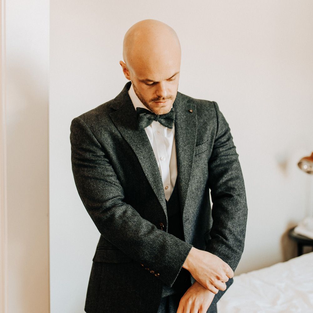 Groom in dark three piece tweed suit with matching bow tie adjusts cuffs as he gets ready for wedding