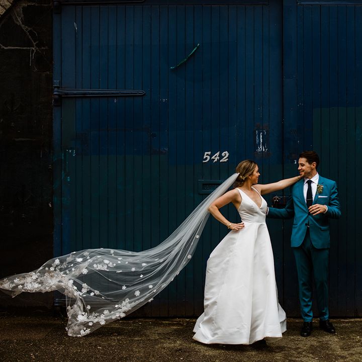 Bride wears long veil with 3D flowers and v neck wedding dress with groom in blue suit 