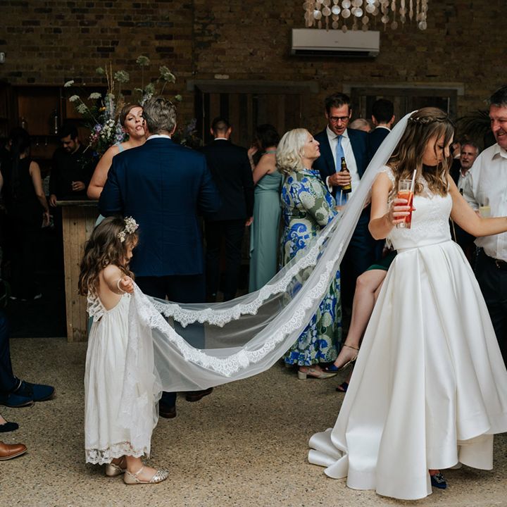 Wedding guests drink and part as flower girl holds up the bride's lace detailed veil 