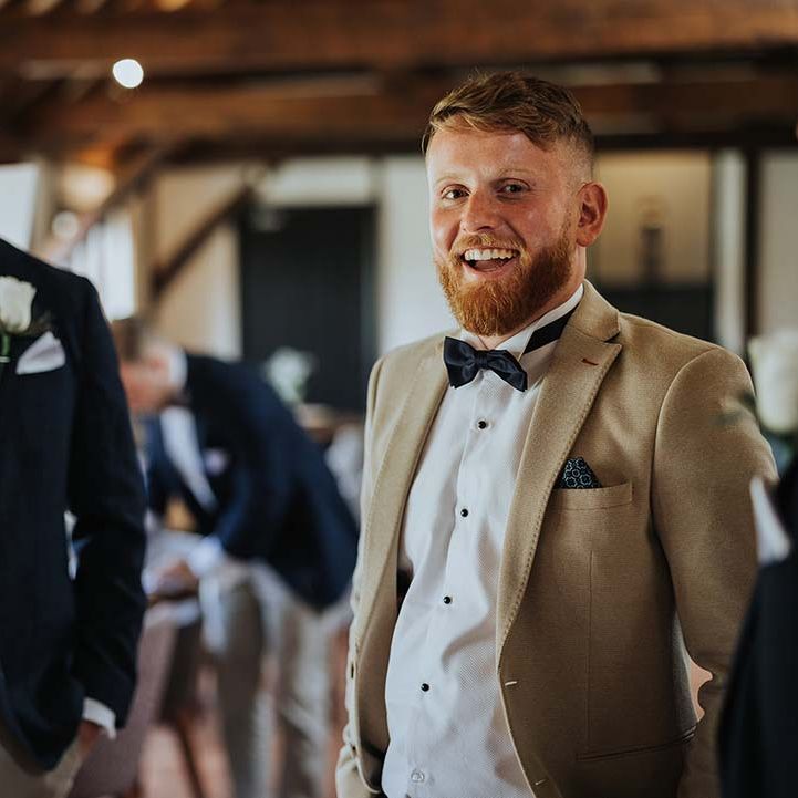 Groom in beige suit jacket and groomsman in dark suit jacket ad burnt orange coloured bow tie for rustic wedding