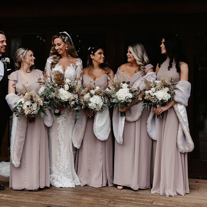 Bride in lace wedding dress standing with bridesmaids in taupe coloured dresses with various neckline styles and matching fur shawls
