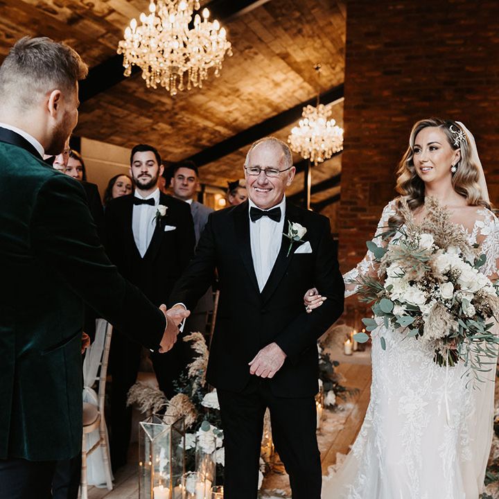 Father of the bride in black tie shakes hands with the groom in green velvet suit jacket as he walks daughter down the aisle