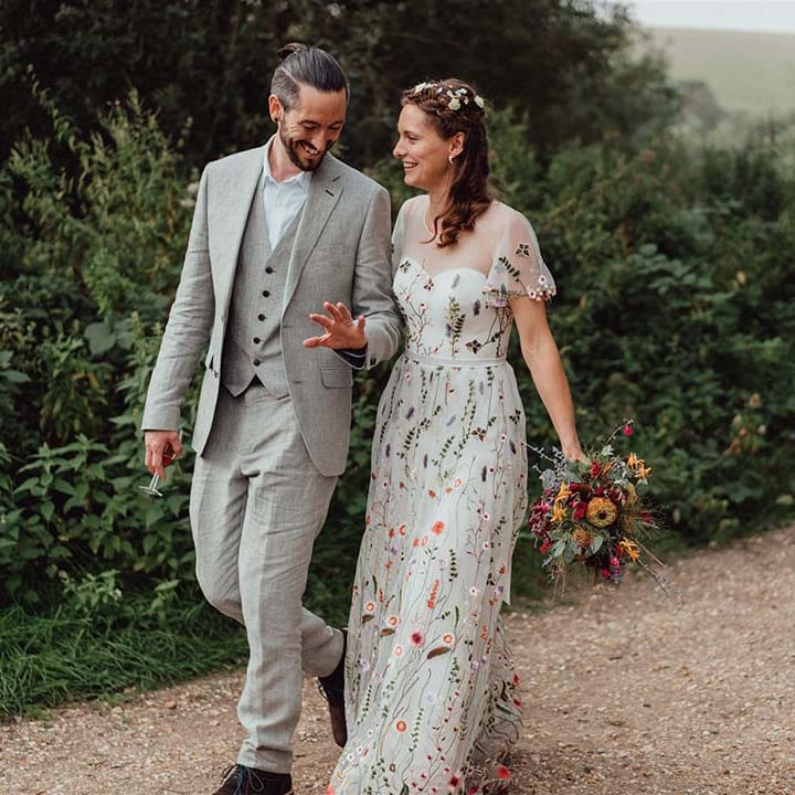 Groom in grey smiles at his wedding ring as he walks with the bride in floral wedding gown and flower hair accessories 