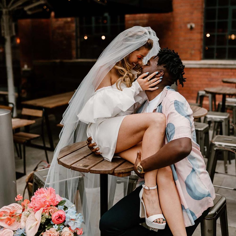 bride in a short wedding Dress and platform shoes sitting on a table with her groom in a patterned shirt embracing her