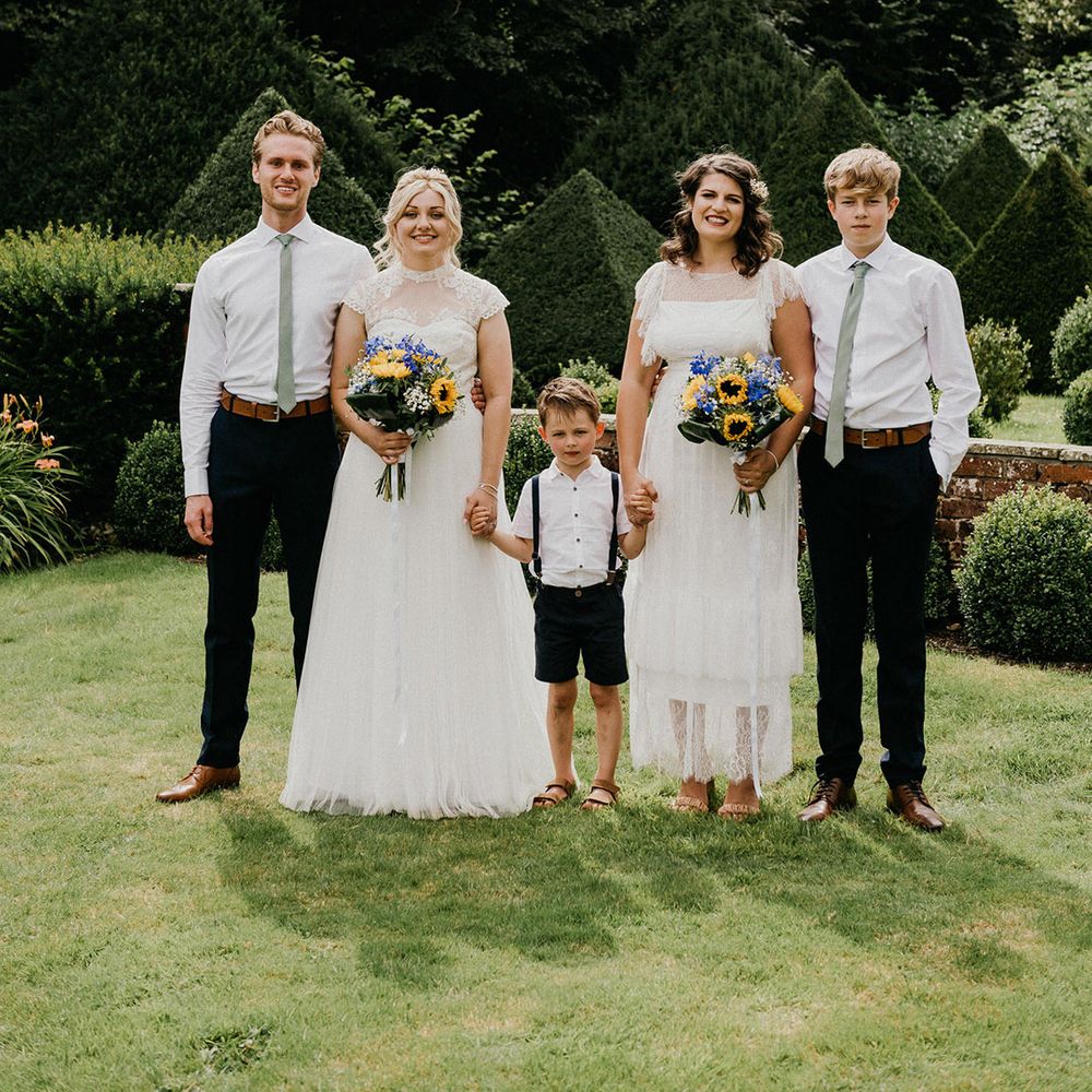 Brides in sheer high neck wedding dresses hold hands with young boy and hold wedding bouquets with sunflowers and gypsophila with older children next to them