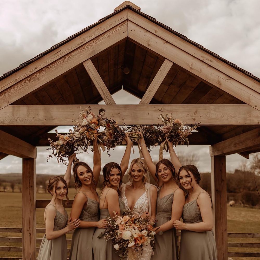 Bridesmaids in green dresses with the bride in lace wedding dress lifting orange and white bouquets 