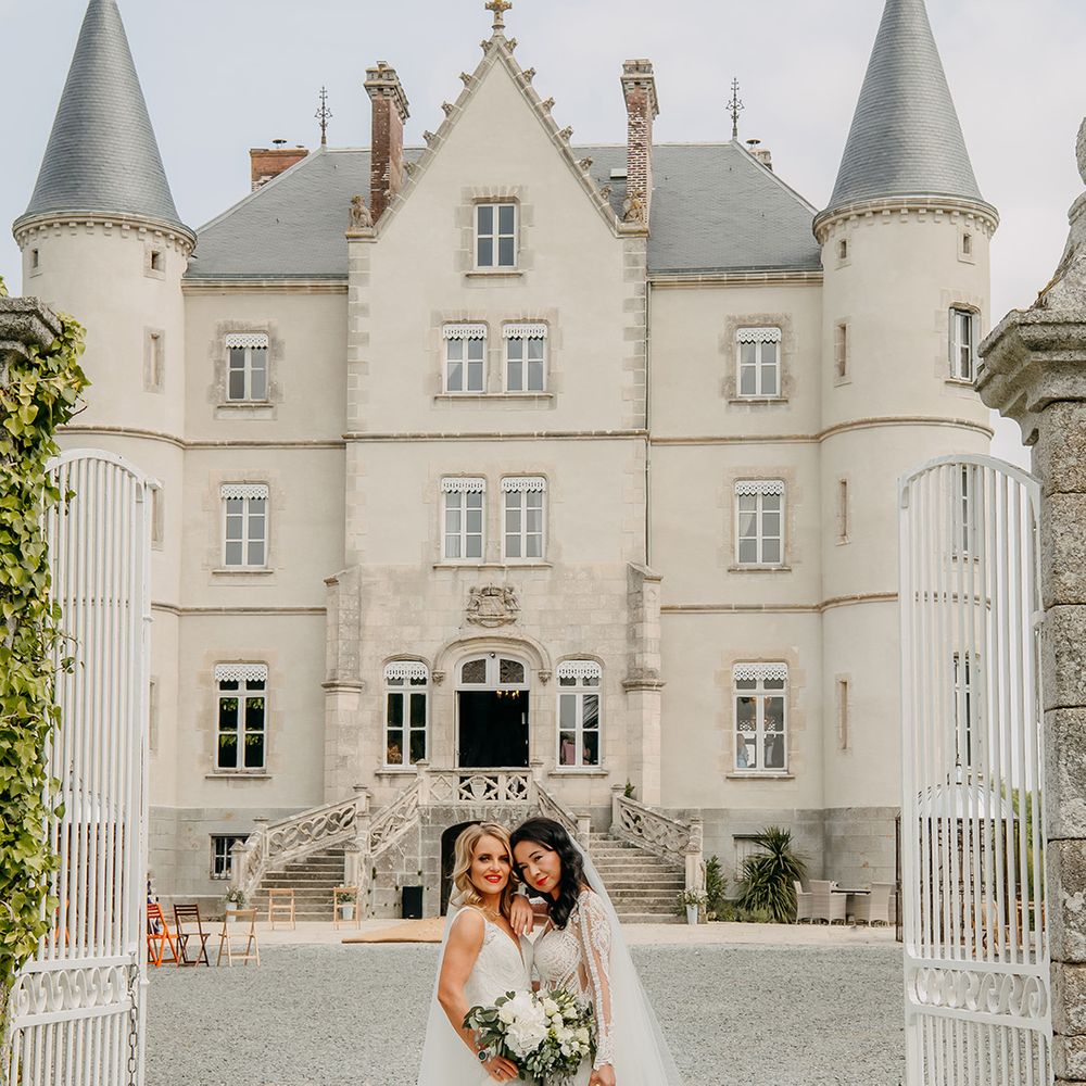 Bride leans against bride with blonde hair as they pose together in their lace wedding dresses