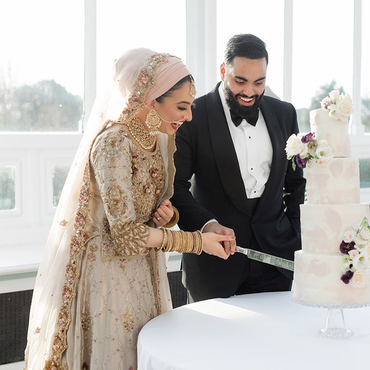Bride & groom cut their four tier wedding cake during reception as groom wears black tie and bride wears traditional saree 