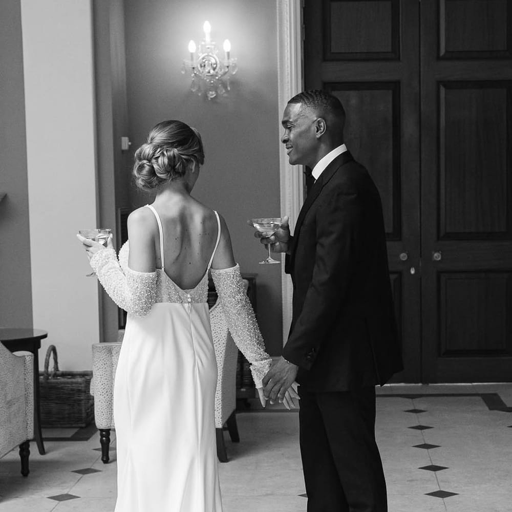 Bride in evening wedding dress with beads and off the shoulder detail walking with groom in black tie as they drink cocktails 