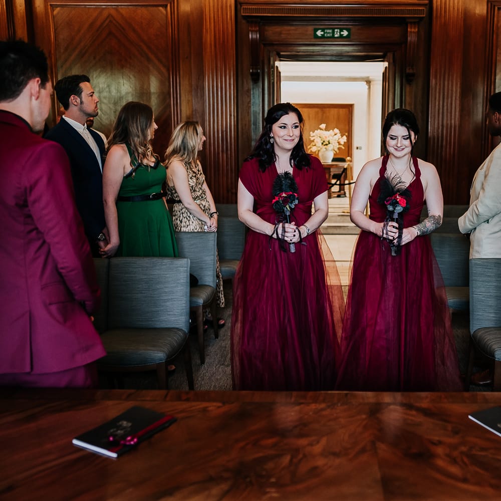 Bridesmaids wear deep red bridesmaid dresses in different styles as they walk down the aisle and hold black & red bouquets complete with black feathers 