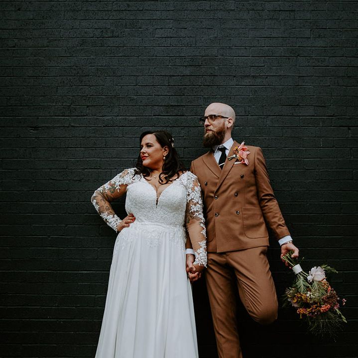 Bride wears lace embellished long sleeve wedding dress whilst stood beside her groom in brown 70s inspired retro suit