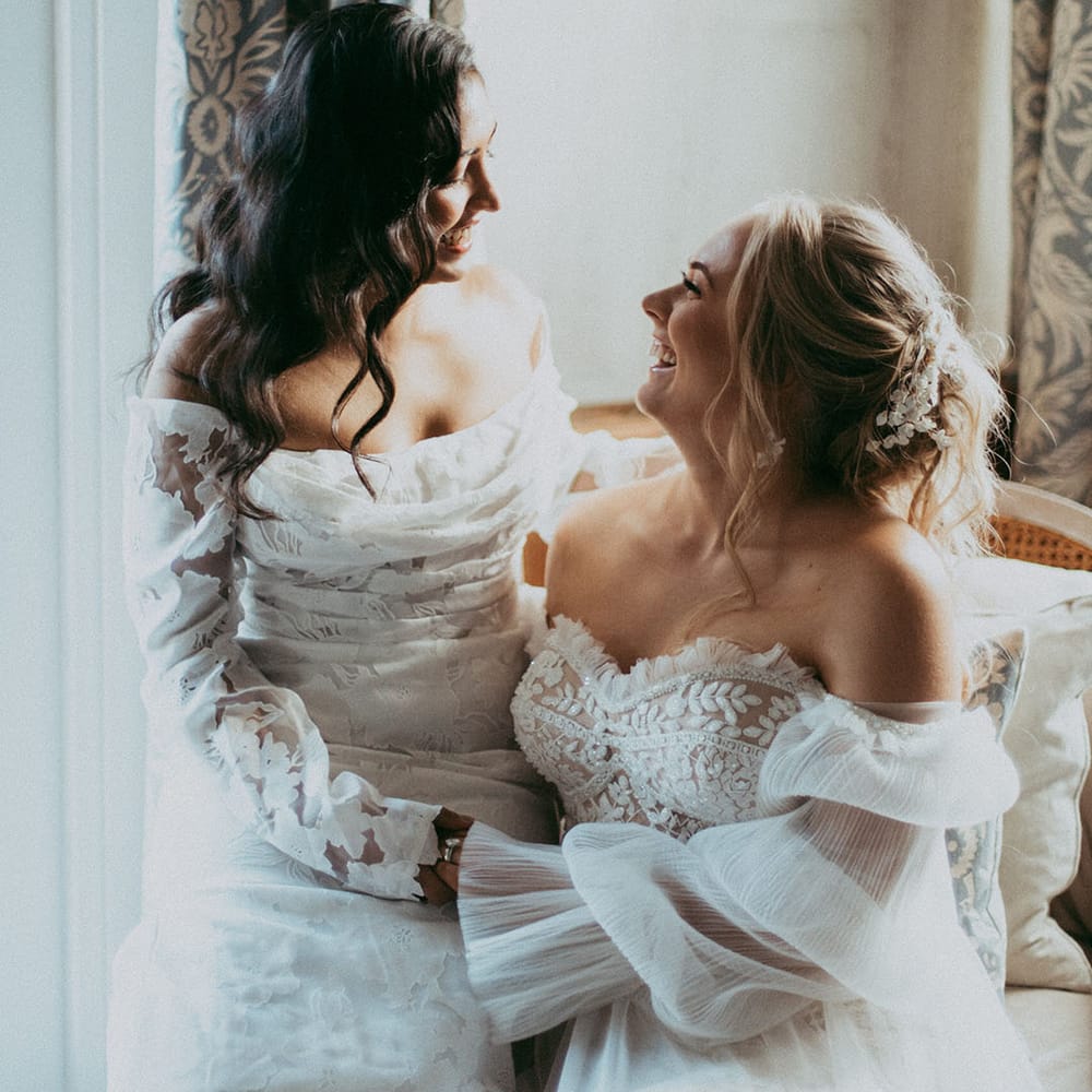 Bride in off the shoulder long sleeve wedding dress sitting on bride's lap wearing strapless lace wedding dress on cream coloured chair, laughing together