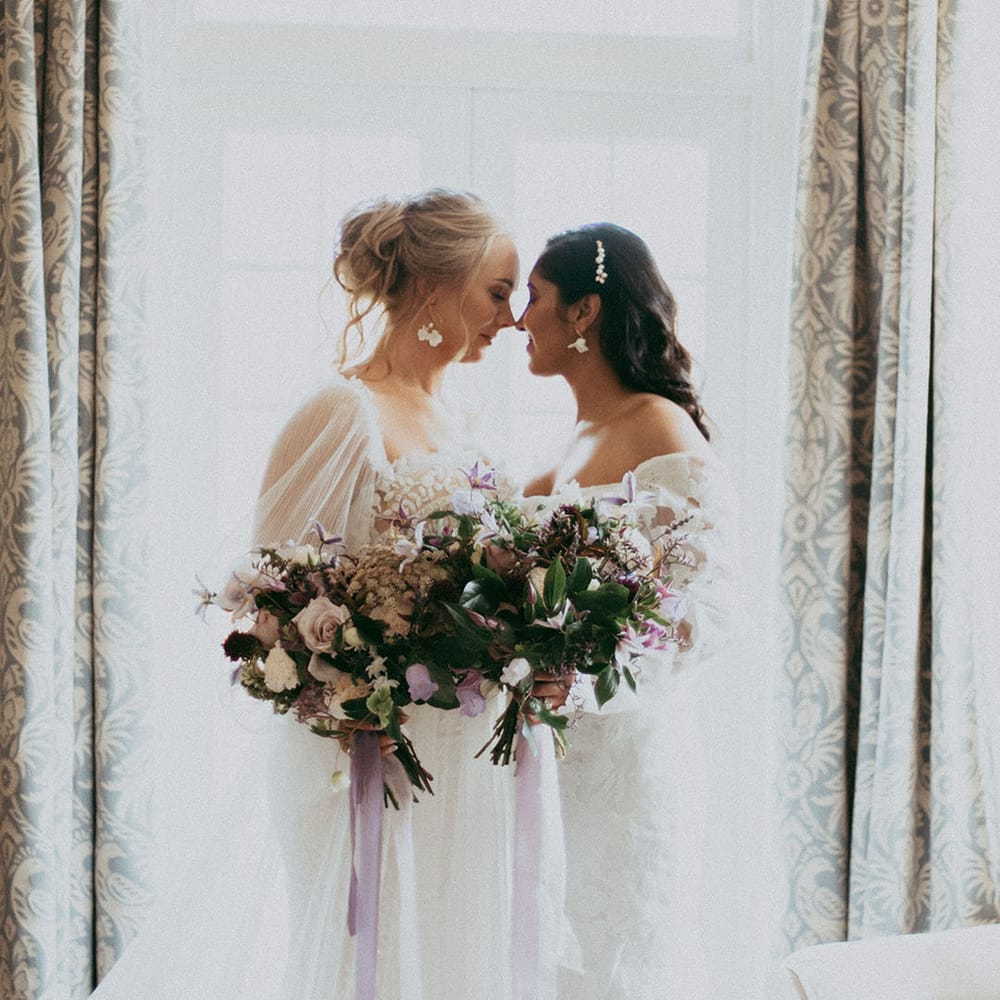 Brides in complimenting lace detail long sleeve wedding dresses holding matching bridal bouquets with ixed neutral floral bouquet with white carnations, purple tulips, roses and foliage
