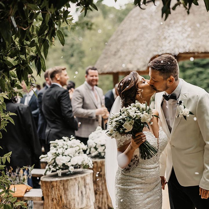 Bride in flower lace wedding dress kisses the groom in a white tux after their confetti exit 