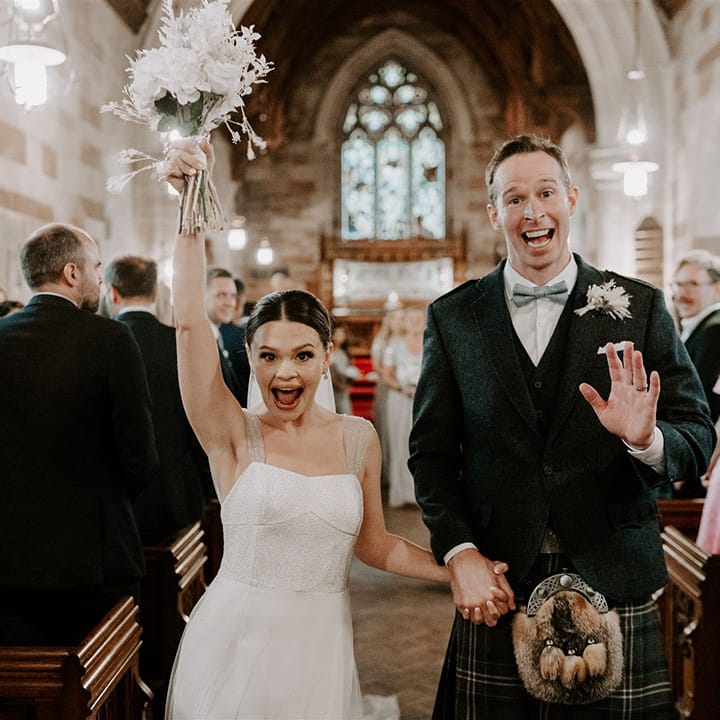 Ecstatic bride lifts up her white bouquet with dried flowers walks out of the church with the beaming groom in a kilt