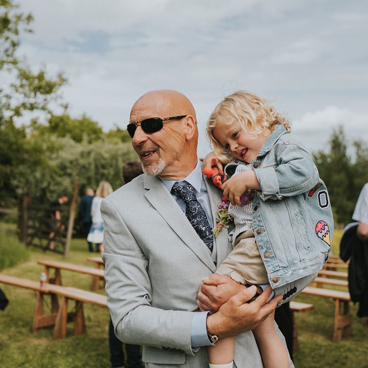 Wedding guest holds little boy in personalised denim jacket during outdoor wedding ceremony 