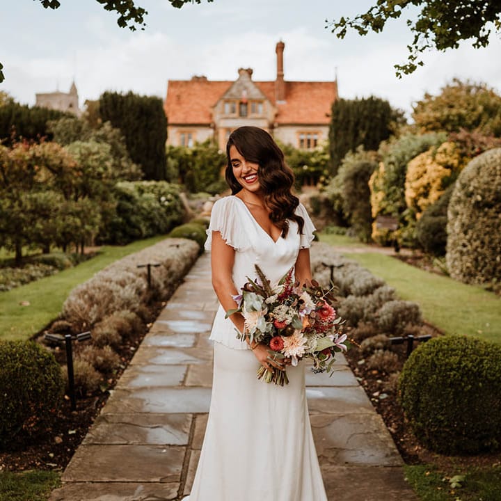 Bride in v neck satin wedding dress with sheer butterfly sleeves and ruffled layer on top holding flower bouquet with roses, carnations, eucalyptus, baby's breath and dried flowers 