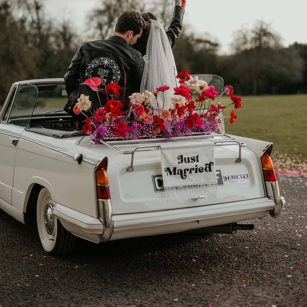 Bride in short wedding veil with delicate flower embroidery at the base and groom in embroidered black leather jacket   “to the moon and back” lettering with crescent moon, stars, and roses embroidery, standing in the back of the off white classic convertible wedding car with red roses, off white peonies, lilac dried lowers and white fabric “just married”  banner decorations