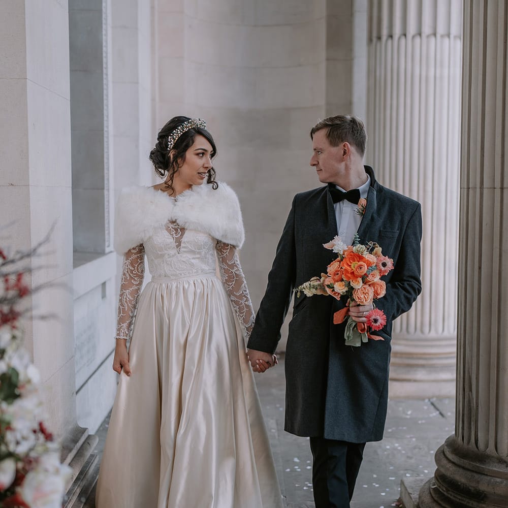 Groom in black overcoat, classic black tuxedo and a black bowtie holding bouquet with light pink poppies, orange garden roses, eucalyptus, Christmas red berry garlands, dried flowers and sage green and orange ribbon with bride in Bride in taffeta material princess wedding dress with long lace sleeves  and cropped faux fur bridal cover up 