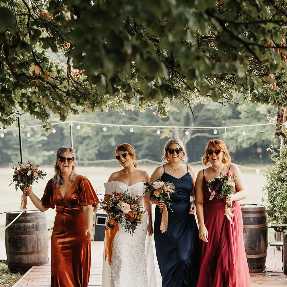 Bride in sunglasses walks alongside her bridesmaids in mismatched bridesmaid dresses in Autumnal colours