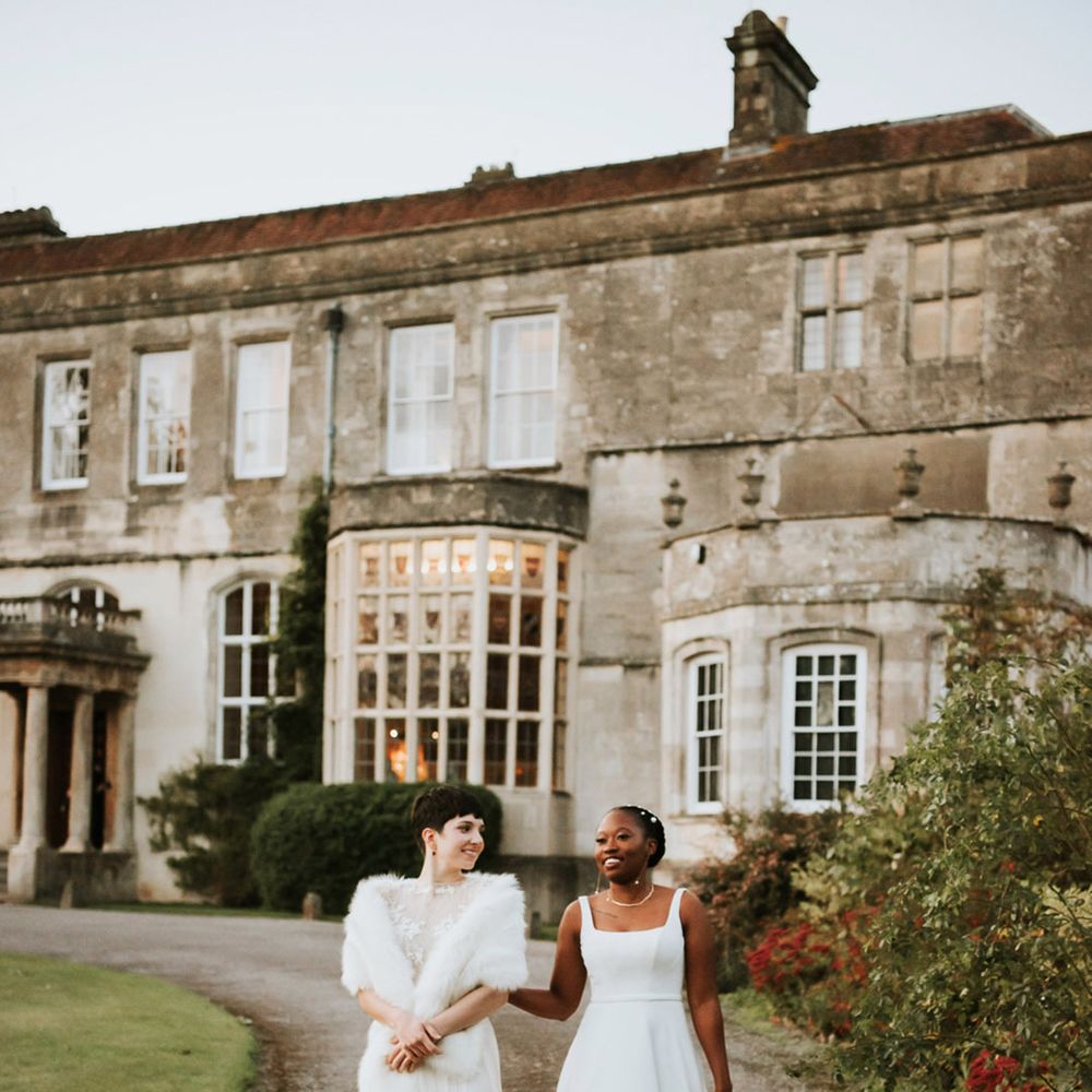 Bride in sleeveless wedding dress walking with bride in short sleeve wedding dress and faux fur shawl walking around the grounds of Elmore Court 