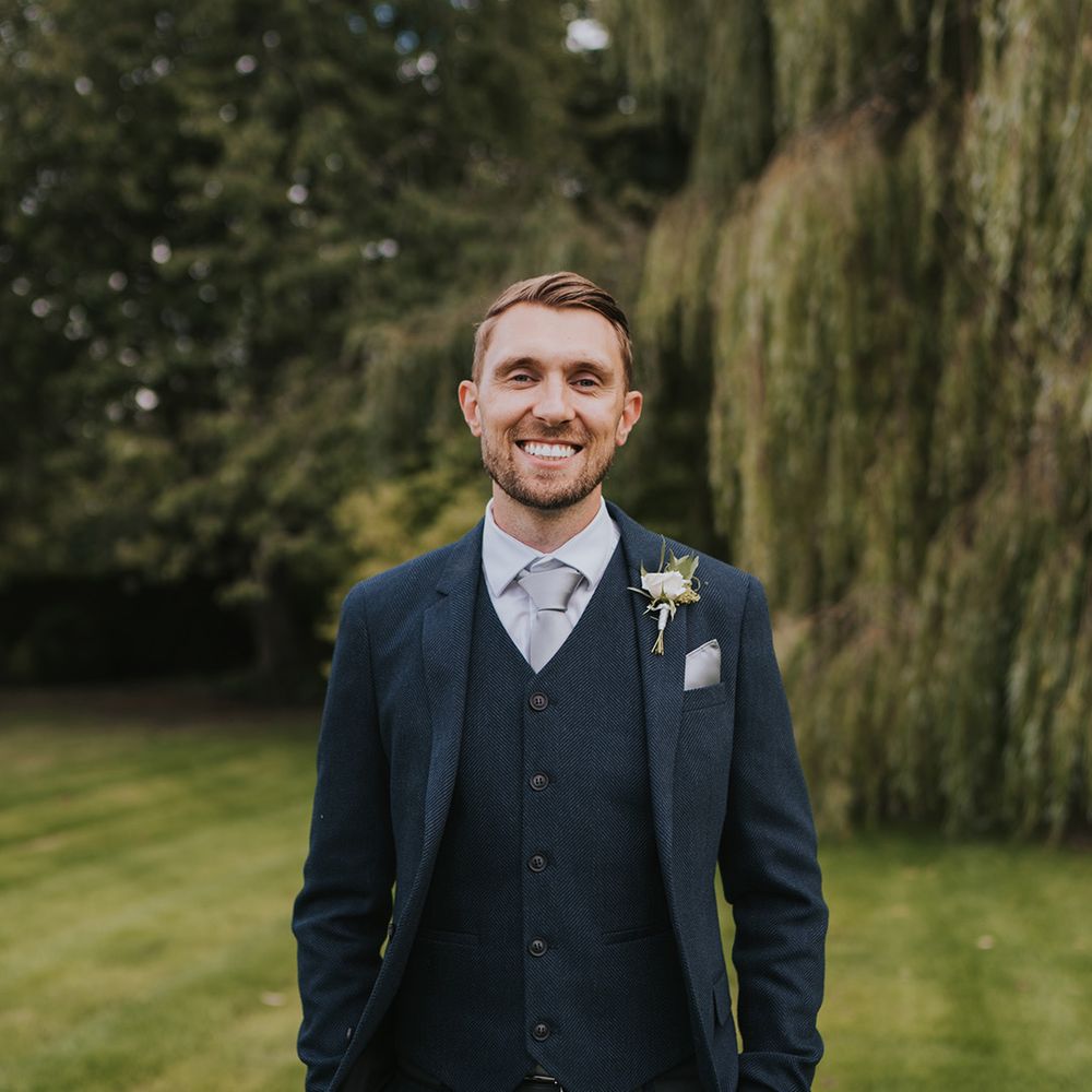 The groom in a three piece dark navy suit with silver grey tie and white rose and gypsophila buttonhole 