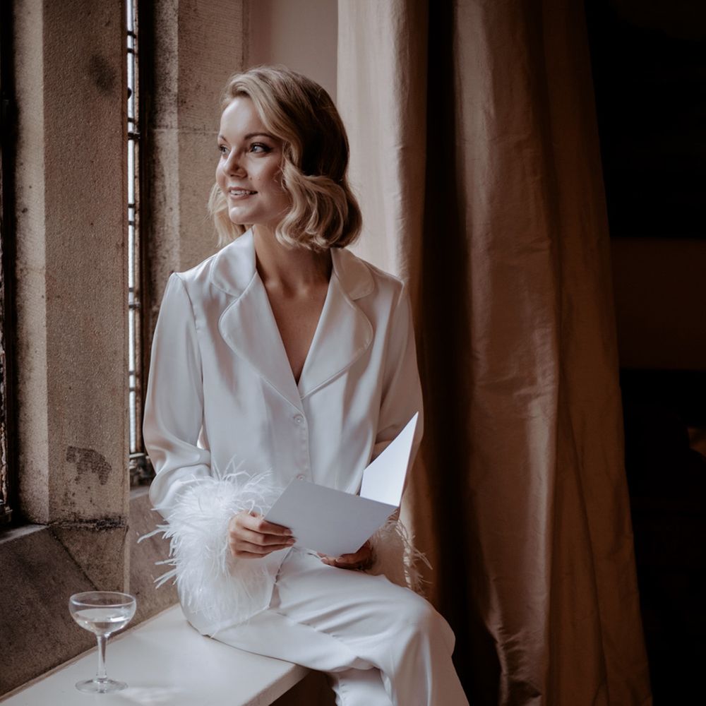Bride in white satin pyjamas with feather cuffs sitting on a window sill reading her wedding vows on the morning of the wedding day 