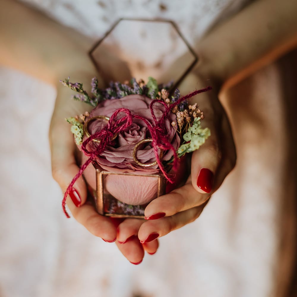 Glass and gold ring box with a dusty pink rose inside and two gold rings tied in red string