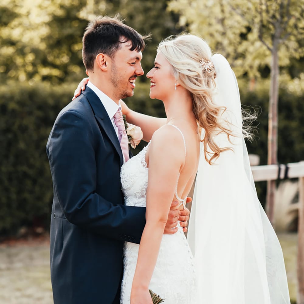 Groom in navy suit with pink tie smiling with the bride in a fitted lace wedding dress with a long veil holding a white wedding bouquet 