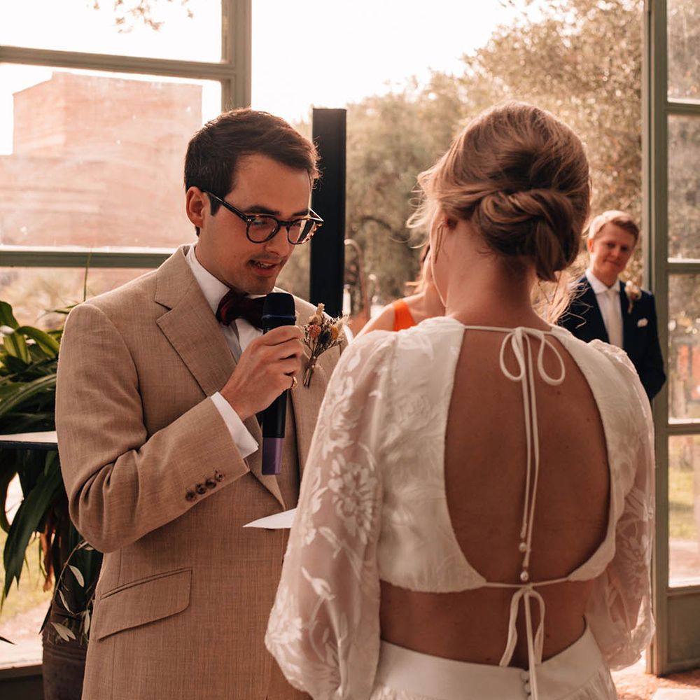 Groom in light cream suit with dried flower buttonhole with tortoise shell glasses reading out his wedding vows to the bride in a Grace Loves Lace bridal two piece for destination wedding 