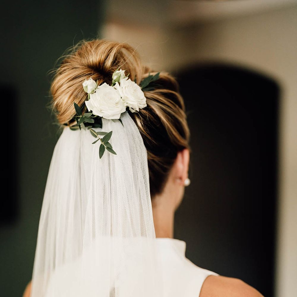 Bride with white rose floral hair accessory attached to bun hairstyle with white veil 