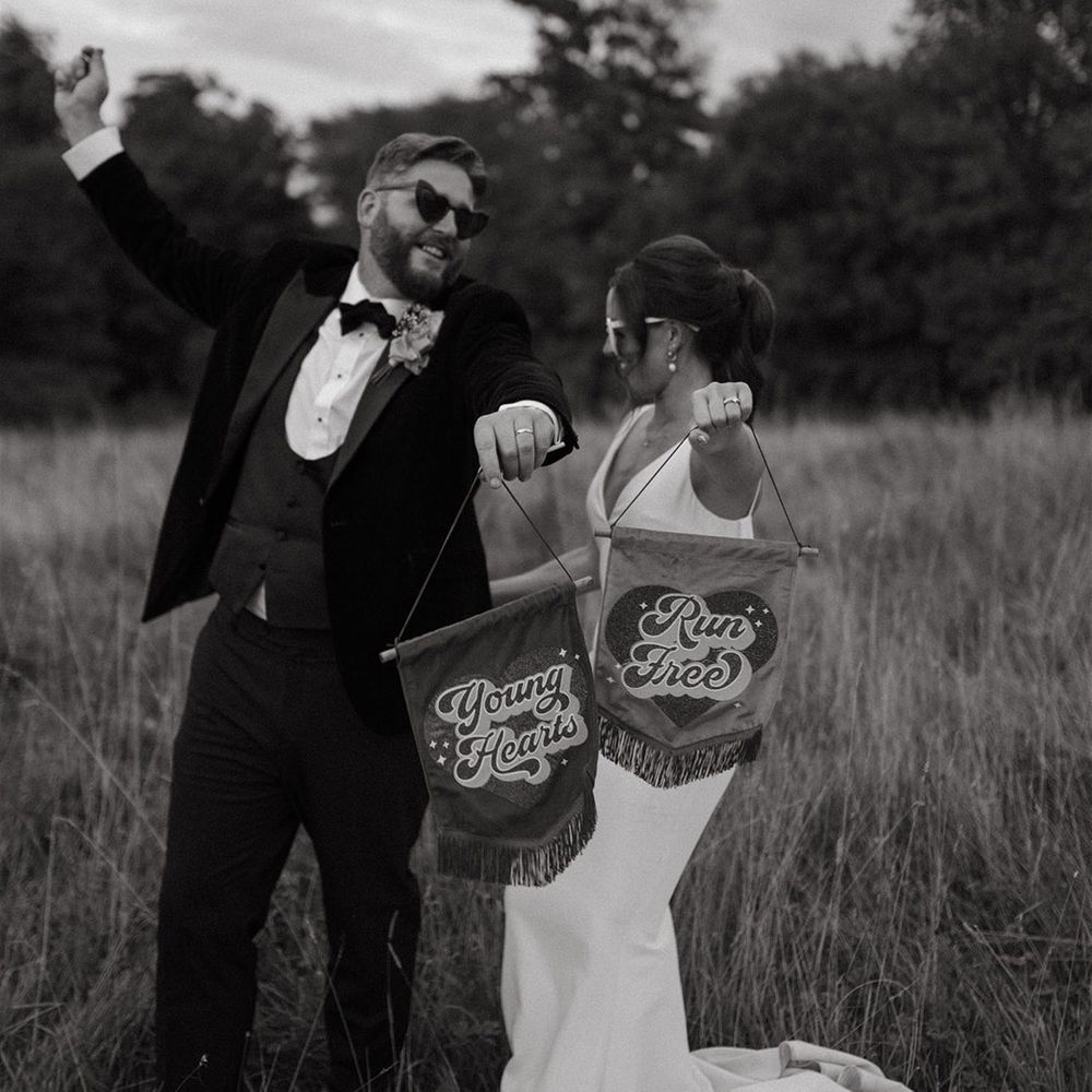 Groom in three piece black tuxedo with the bride in a satin slip dress holding pink and red retro banner signs 
