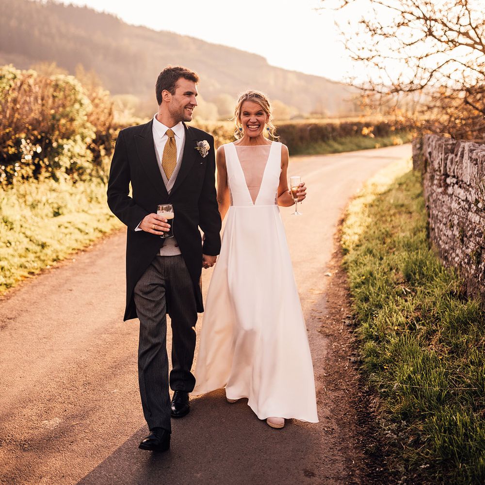 Groom in morning suit with yellow tie holding glass of Guinness walking with bride in Andrea Hawke wedding dress 