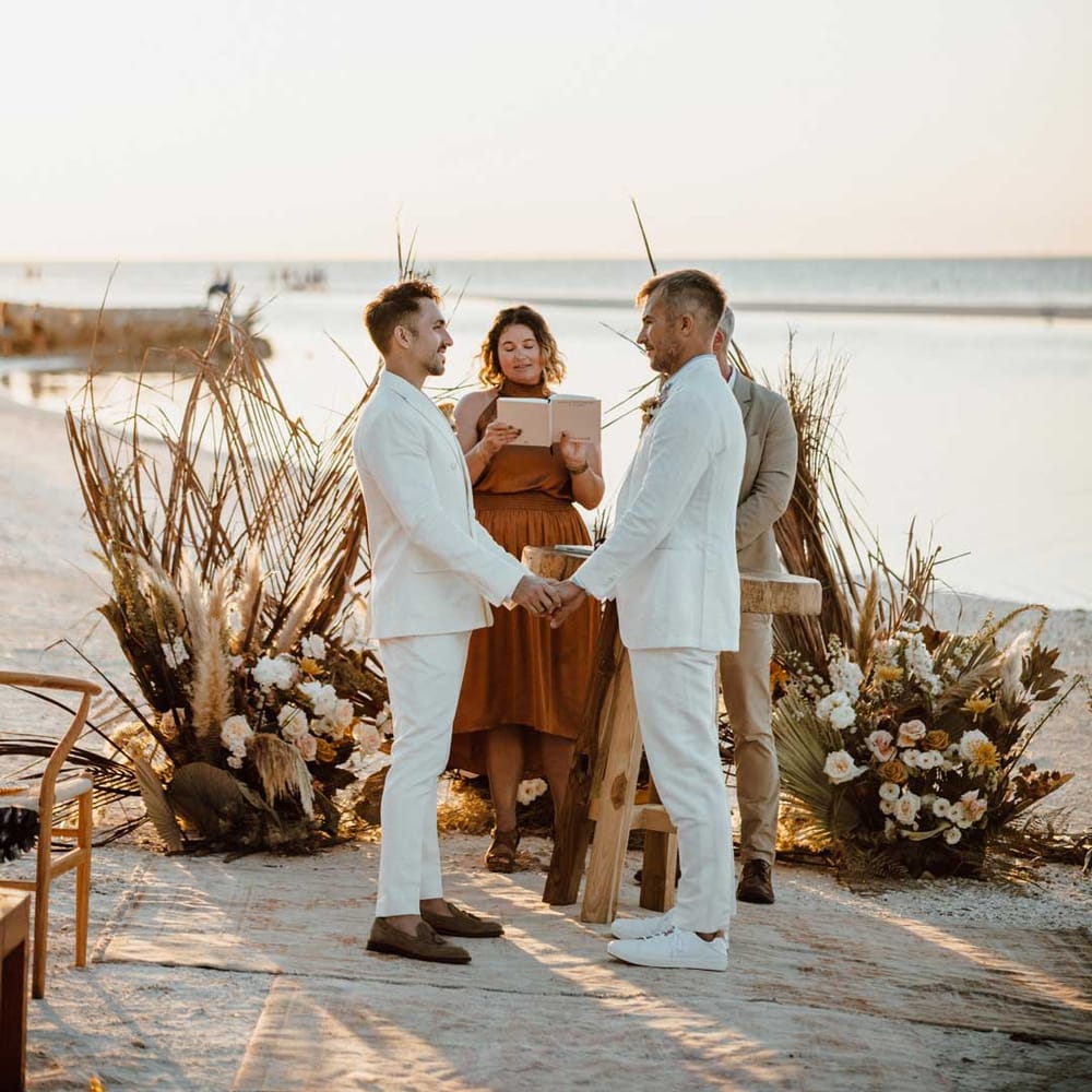 Two grooms in white suits hold hands for their LGBTQI+ wedding ceremony at beach wedding 