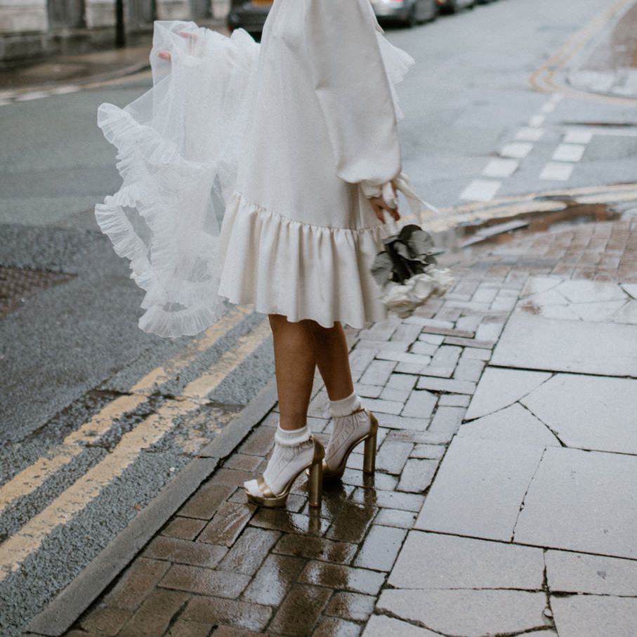 Bride in a silk wedding dress, socks and gold Gucci platform shoes holding her veil by the side of the road 