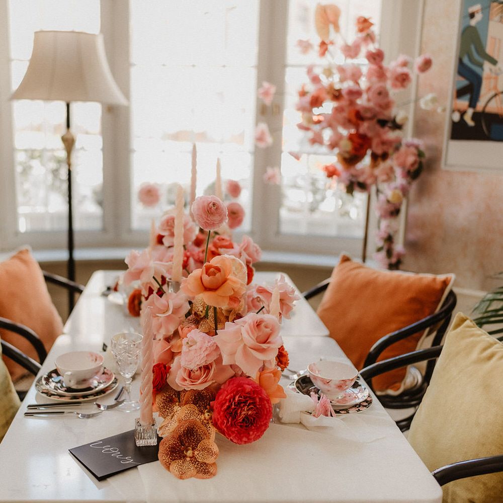 Luxurious table setup with tapered candles, floral teacup and & saucers, velvet seating and peach wedding flowers including Pink, peach and burgundy ranunculi, garden roses and orchids