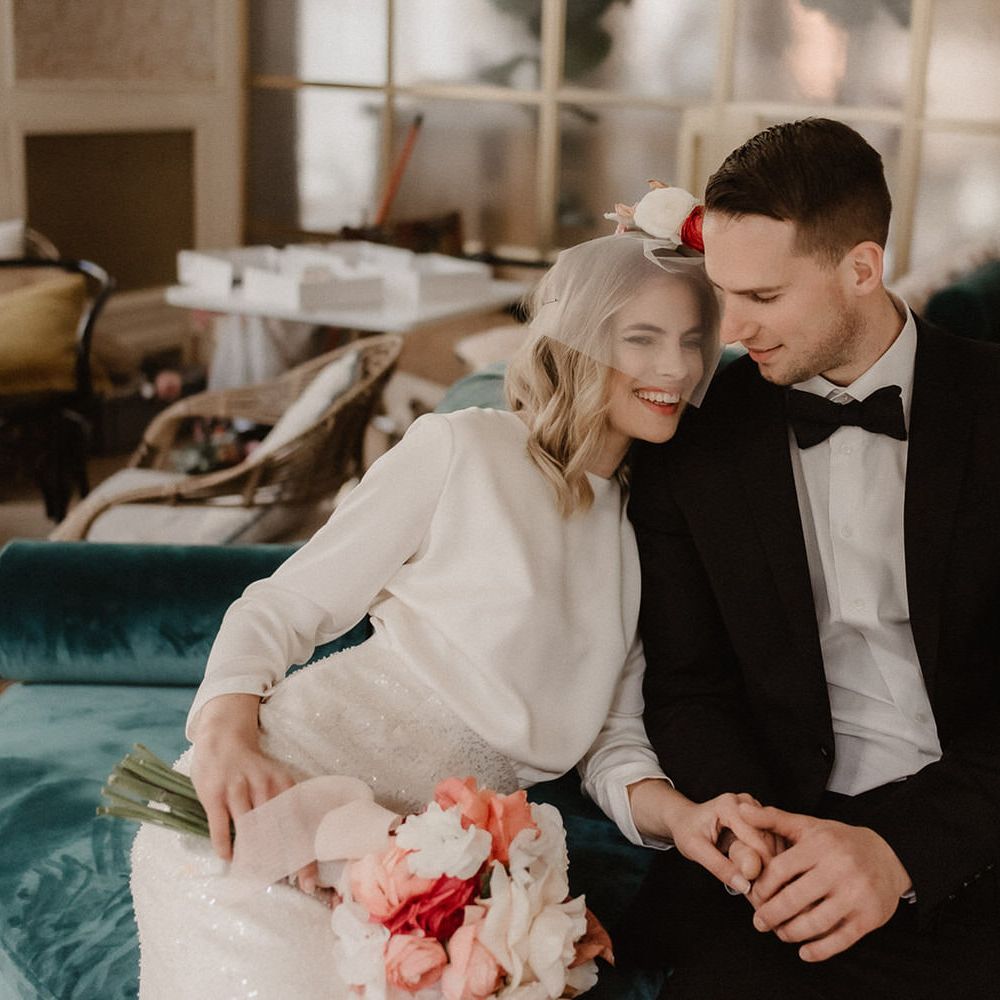 Bride and groom holding hands on a blue velvet sofa, bride is holding a bouquet of pink and white flowers