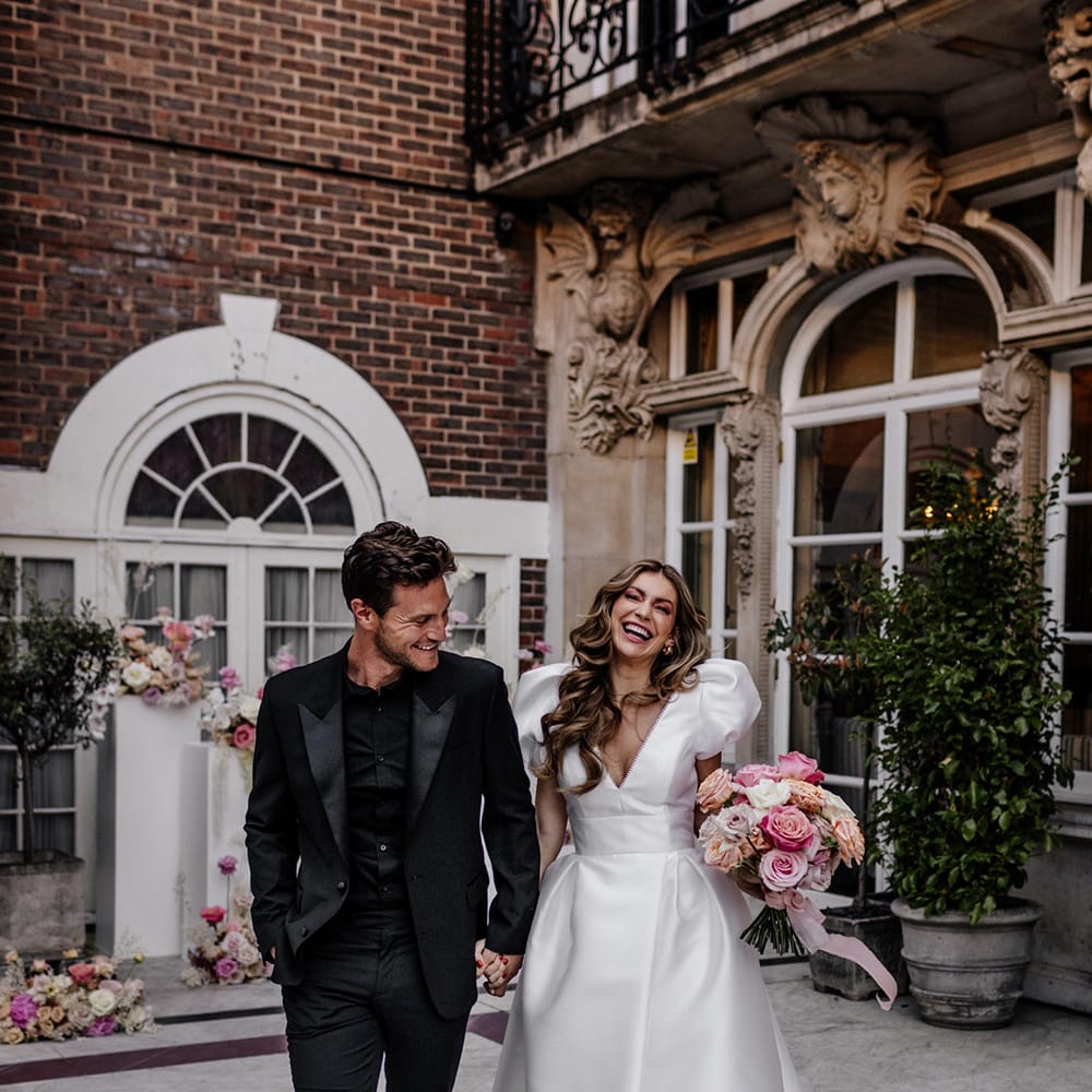 Happy bride and groom in an all black suit, with black shirt and loafers and Bride in a Love in Lace bridal gown with puff sleeves, plunging neckline and open front skirt holding hands 
