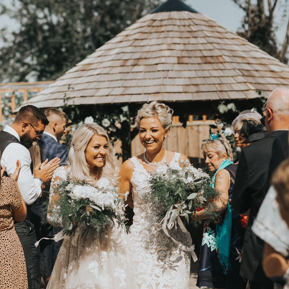 Two brides exit their outdoor wedding ceremony laughing. 