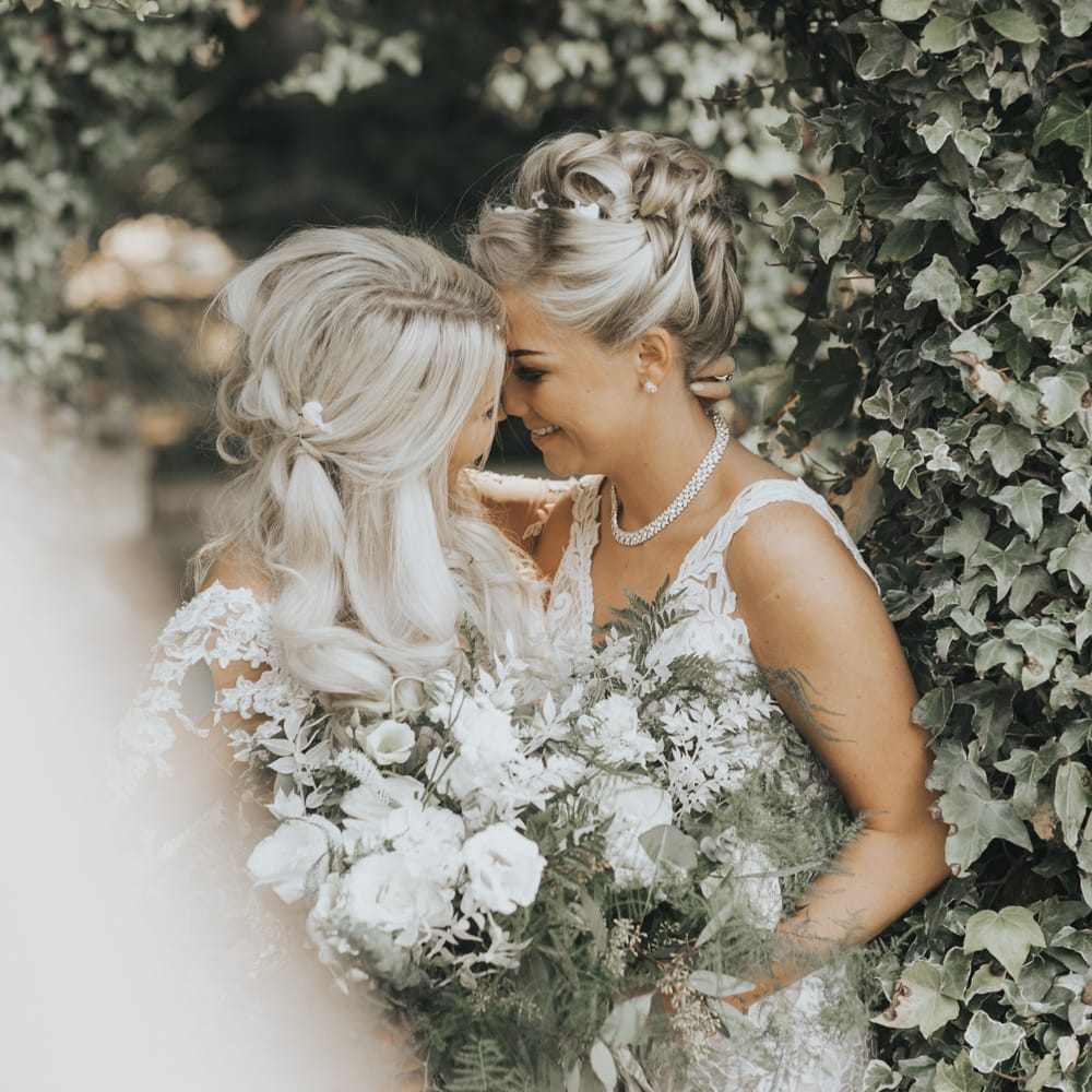 Two brides embrace under a canopy of flowers.
