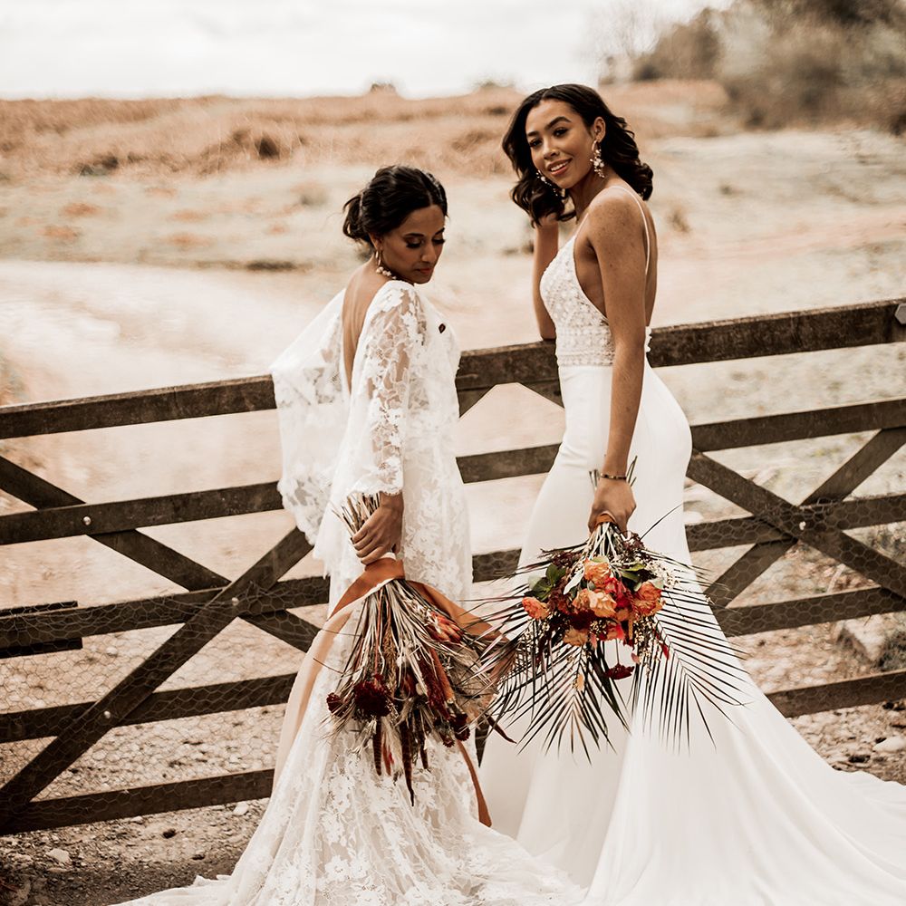 Two bride in, lace and fitted wedding dress holding orange bouquets standing at a field gate 