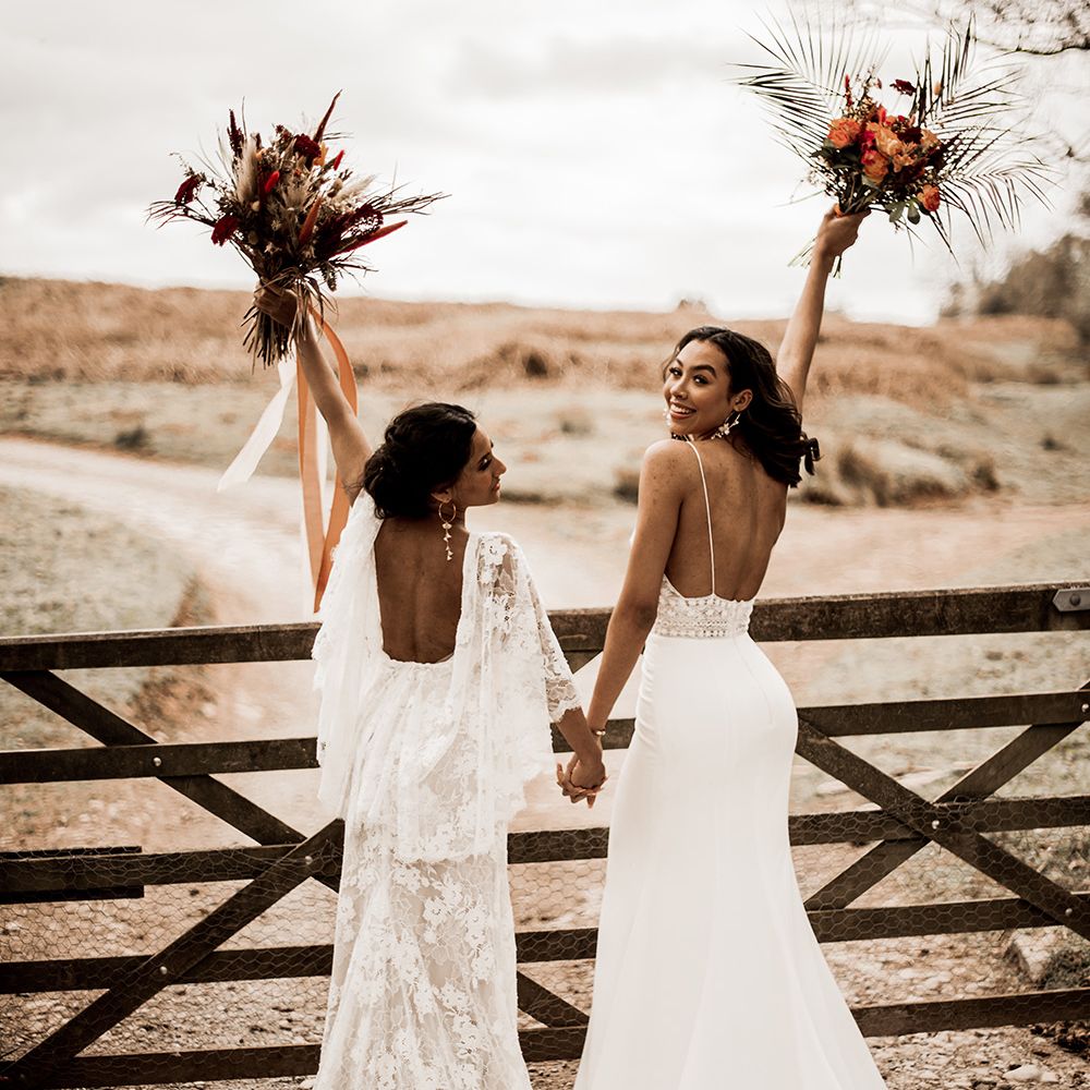 Two brides in a lace wedding dress and fitted wedding dress holding hands and their red bouquets in the air in front of a wooden gate at their farm wedding venue 