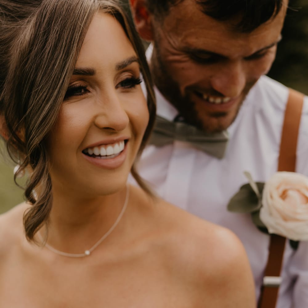 Bride laughs as her groom stands behind her wearing white shirt and white floral buttonhole | Mark Bamforth Photography