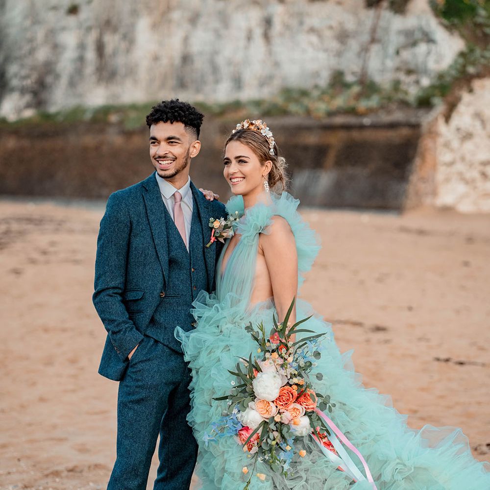 Bride and groom portrait at their coastal elopement with bride in a mint green wedding dress holding a pastel wedding bouquet tied with ribbon and groom in a blue wool suit 
