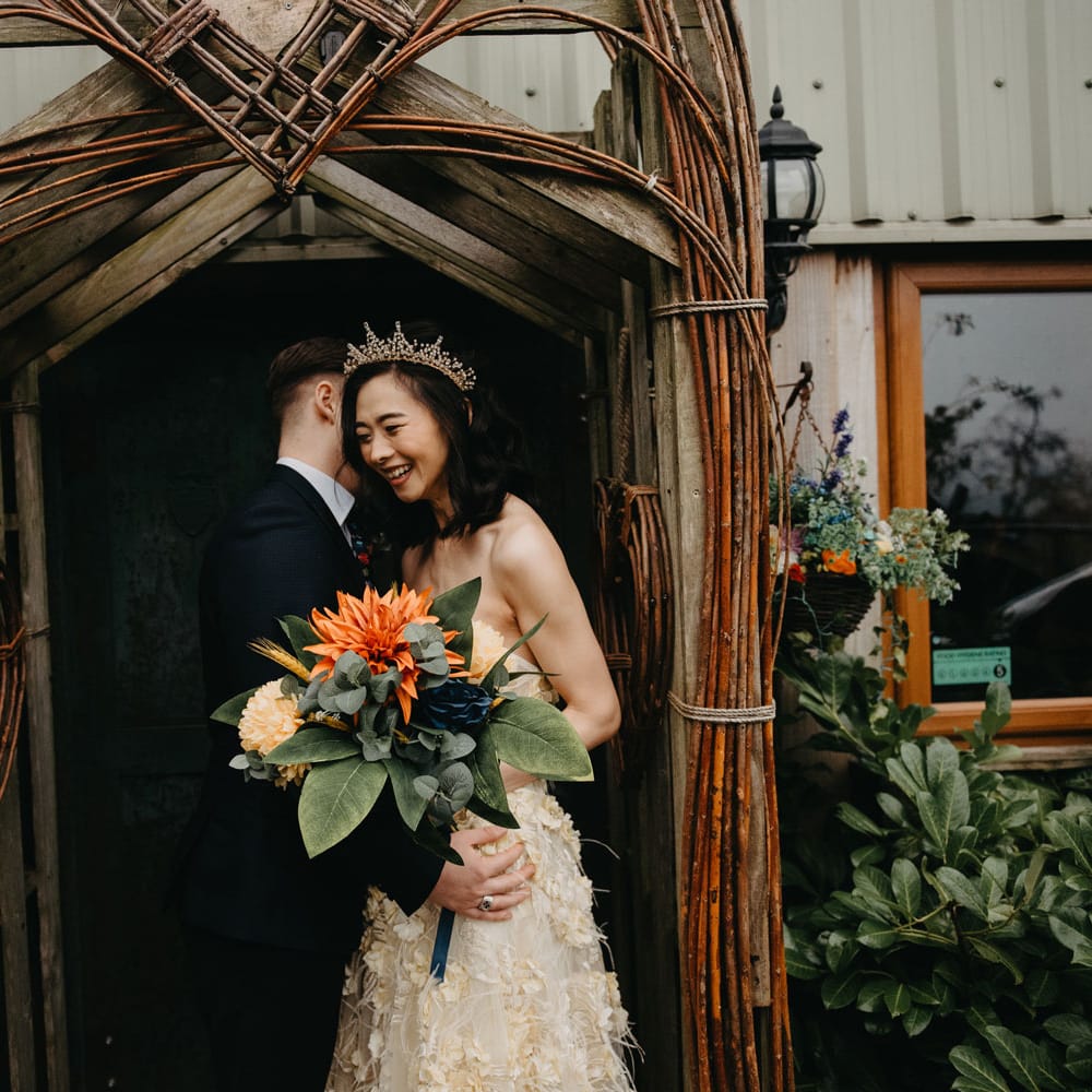 Laughing bride in strapless feathered wedding dress and tropical wedding bouquet stands with groom in dark suit in doorway for farm wedding in Lancashire