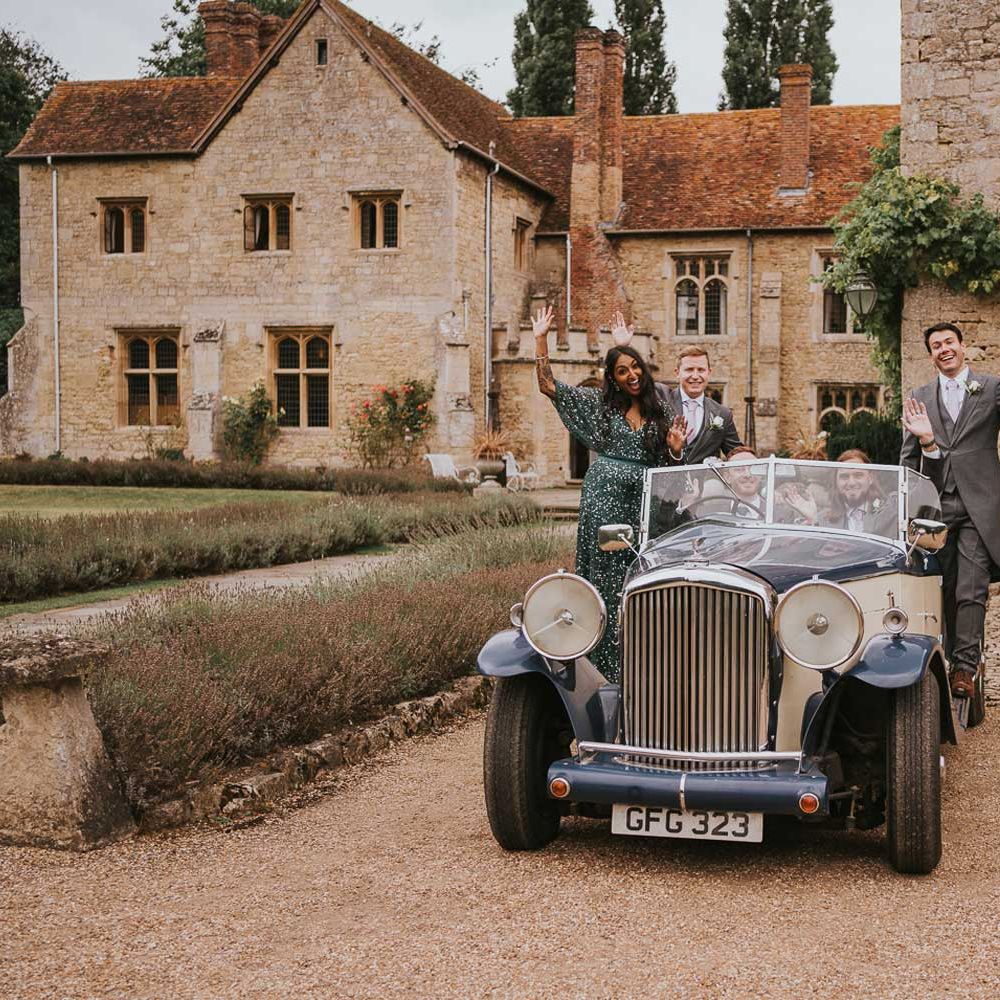 Groom, groomsmen and woman in sequin green dress wave from vintage car parked in the grounds of Notley Abbey in Buckinghamshire 
