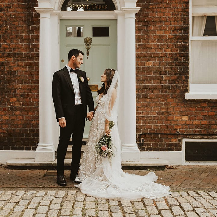 Bride looks up at her groom on her wedding day as they stand in front of pastel green door 
